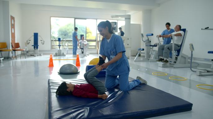 年轻的物理治疗师在物理康复中心帮助一个躺在垫子上的小男孩伸展双腿，而其他患者和治疗师则在后台工作