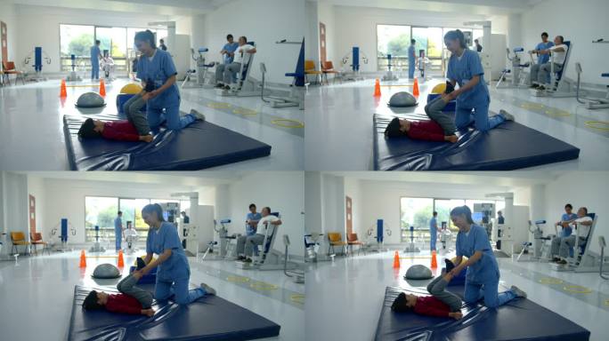 年轻的物理治疗师在物理康复中心帮助一个躺在垫子上的小男孩伸展双腿，而其他患者和治疗师则在后台工作