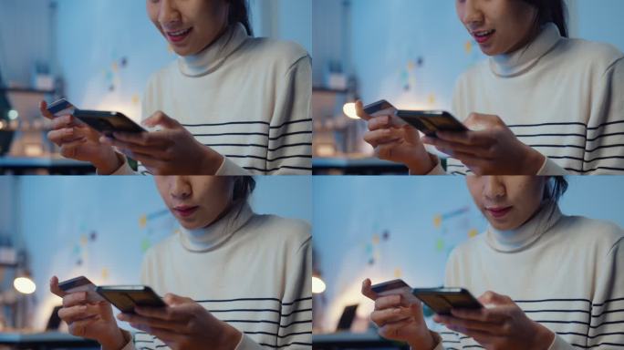 年轻的亚洲女性在晚上坐在家里的沙发上购物时，一边使用手机一边用信用卡进行网上交易。