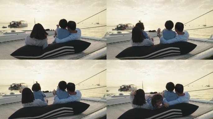 日落时，亚洲家庭坐在游艇上，坐在蹦床上，拥抱在一起，享受着在海上移动的乐趣。年轻的父母和小儿子在夏日