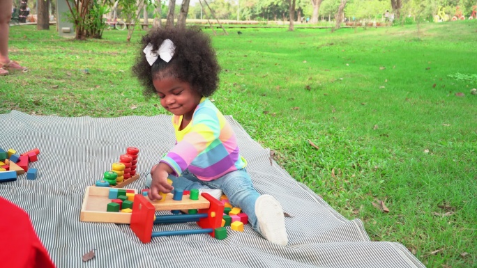 美国一个非洲小女孩，穿着鲜艳的布料，牛仔裤和大发髻，坐在绿色公园草地上的垫子上野餐，用锤子敲打积木玩