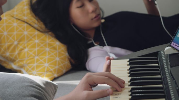 一个亚洲男孩在家里玩手机游戏的妹妹旁边的沙发上，通过手机应用程序学习键盘乐器。