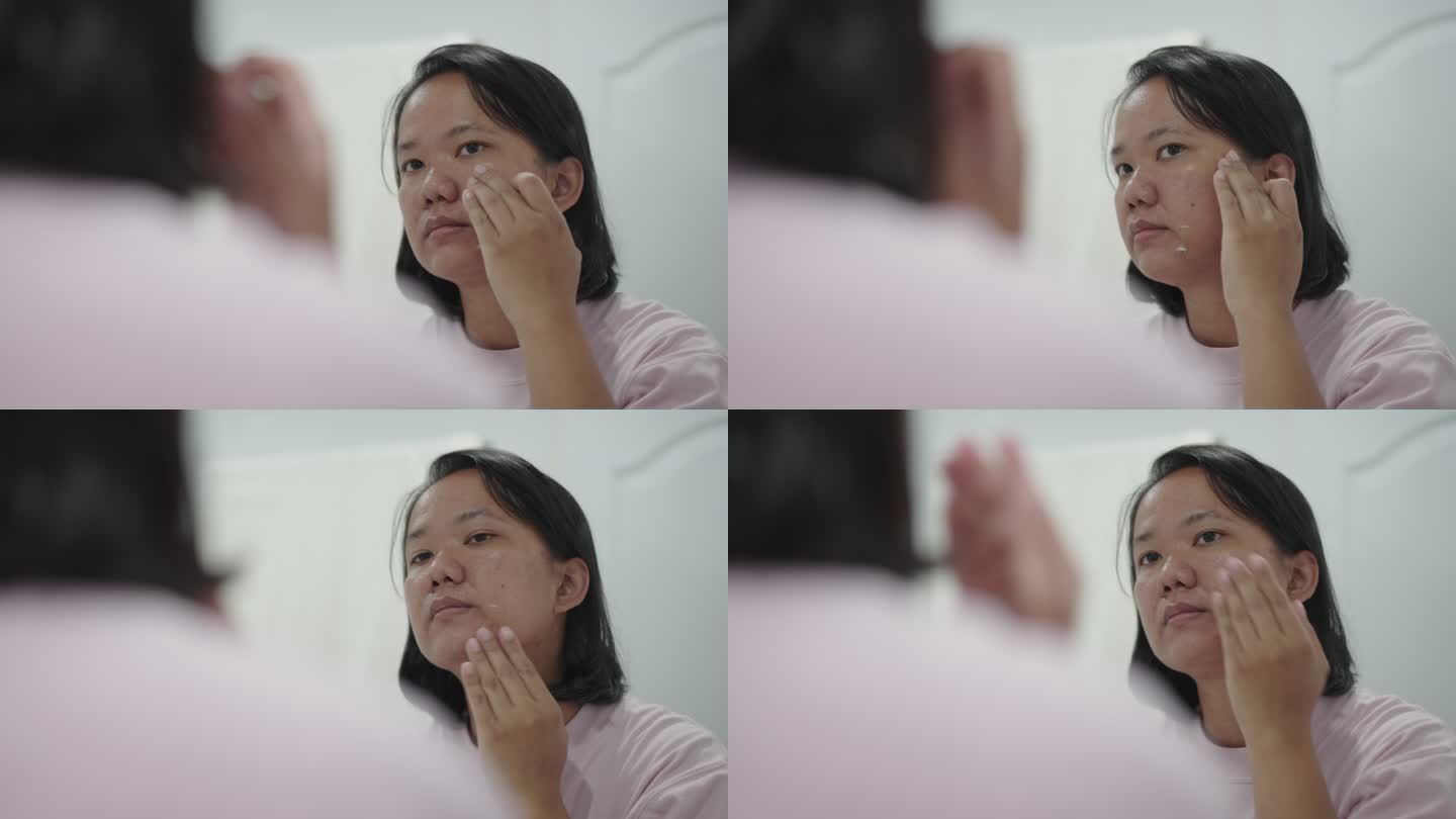 亚洲女人在镜子中看着自己的脸，用面霜治疗