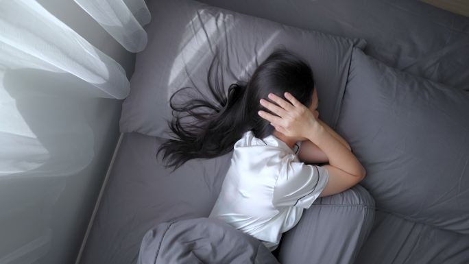 一位亚洲女性因噪音问题而烦躁不安，她试图捂住躺在床上的耳朵睡觉。俯视图。