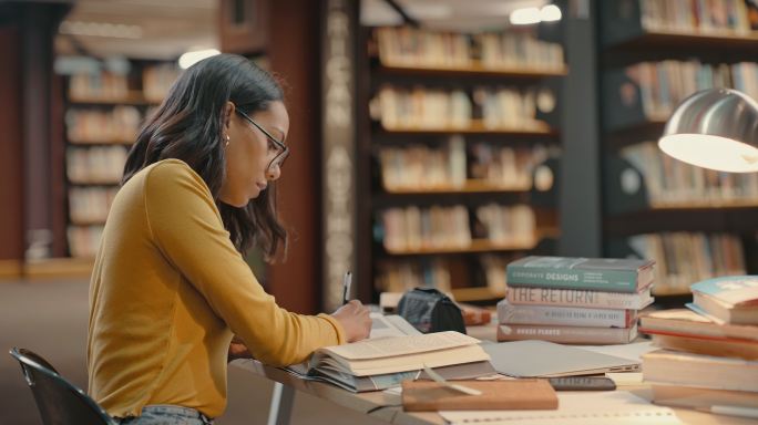 一名学生在学校图书馆备考或为一个项目做研究。一位戴着眼镜的快乐年轻律师坐在书桌旁，手里拿着一堆书，一