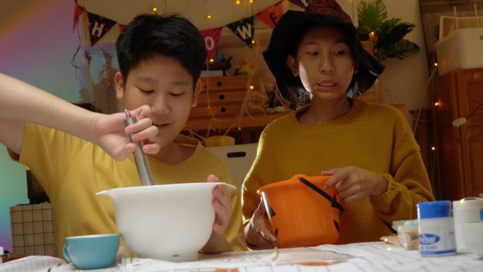 一个亚洲女孩戴着女巫帽，晚上和哥哥一起在家里做糖衣饼干，这是万圣节的生活理念。