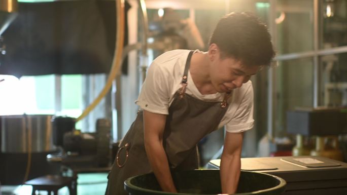 亚裔中国男性工人将生咖啡豆从桶中舀到称重机中，并在工厂仓库中混合用于咖啡烘焙过程