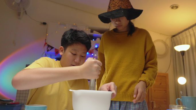 一个亚洲女孩戴着女巫帽，晚上和哥哥一起在家里做糖衣饼干，这是万圣节的生活理念。