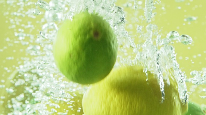 在绿色的工作室背景下，柠檬和酸橙被扔进汽水中。成熟、健康、多汁的水果在液体或液体中游泳，制成柠檬水或