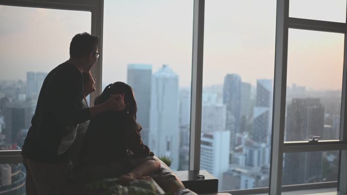 吉隆坡市起居室里，两个孩子透过窗户观看美丽的日落，侧影后视亚裔华人家庭