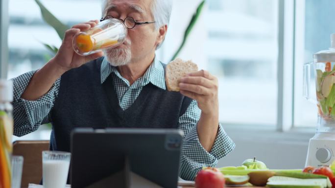 亚洲男性老年白发老人一边吃健康的早餐面包，一边喝水，一边阅读平板电脑上的新闻社会问题