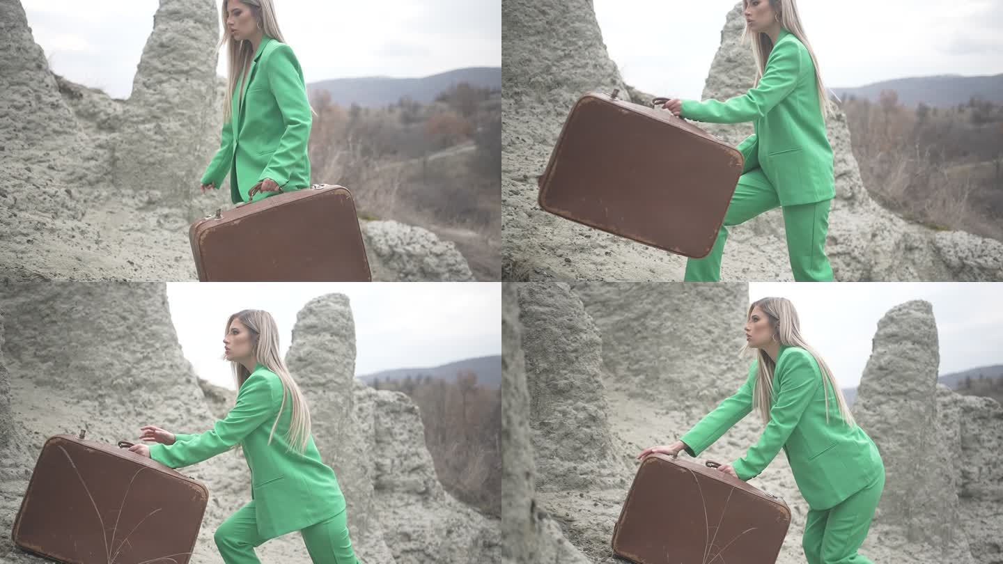 身穿绿色西装的女性拖着沉重的行李箱走向岩石山顶