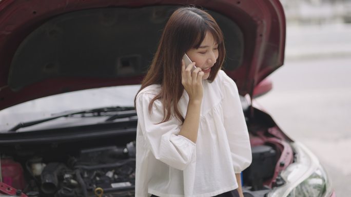 当汽车抛锚时，沮丧的年轻女子打开汽车引擎盖，用智能手机呼救。