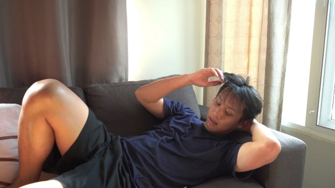 亚洲男人躺在沙发上，在家里用体温计测量体温