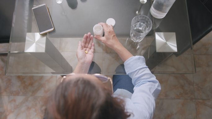 俯视图：正在服药或药丸的亚洲资深女性。一位老妇人坐在家里的餐桌上，吃了一片药丸后，手里拿着水喝着。