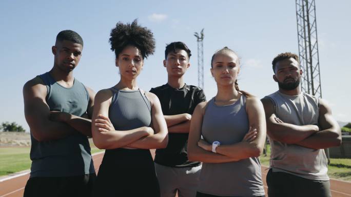 严肃的年轻运动员双臂交叉站在户外田径跑道上的肖像。由自信、积极、专注的运动男女组成的多元化团队，准备