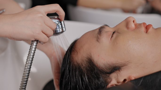 美发师正在用水清洗客户亚洲男性的头发。一个年轻人闭着眼睛在发廊洗头。