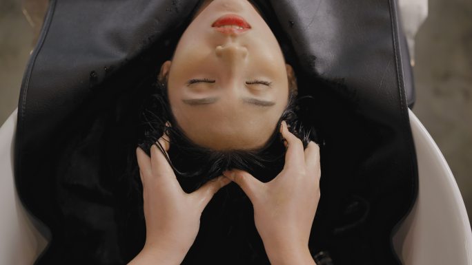一位美丽的亚洲女孩闭着眼睛在美容院理发前躺下洗头。美发师的女性手在美容院洗头时按摩客户的头部。造型师