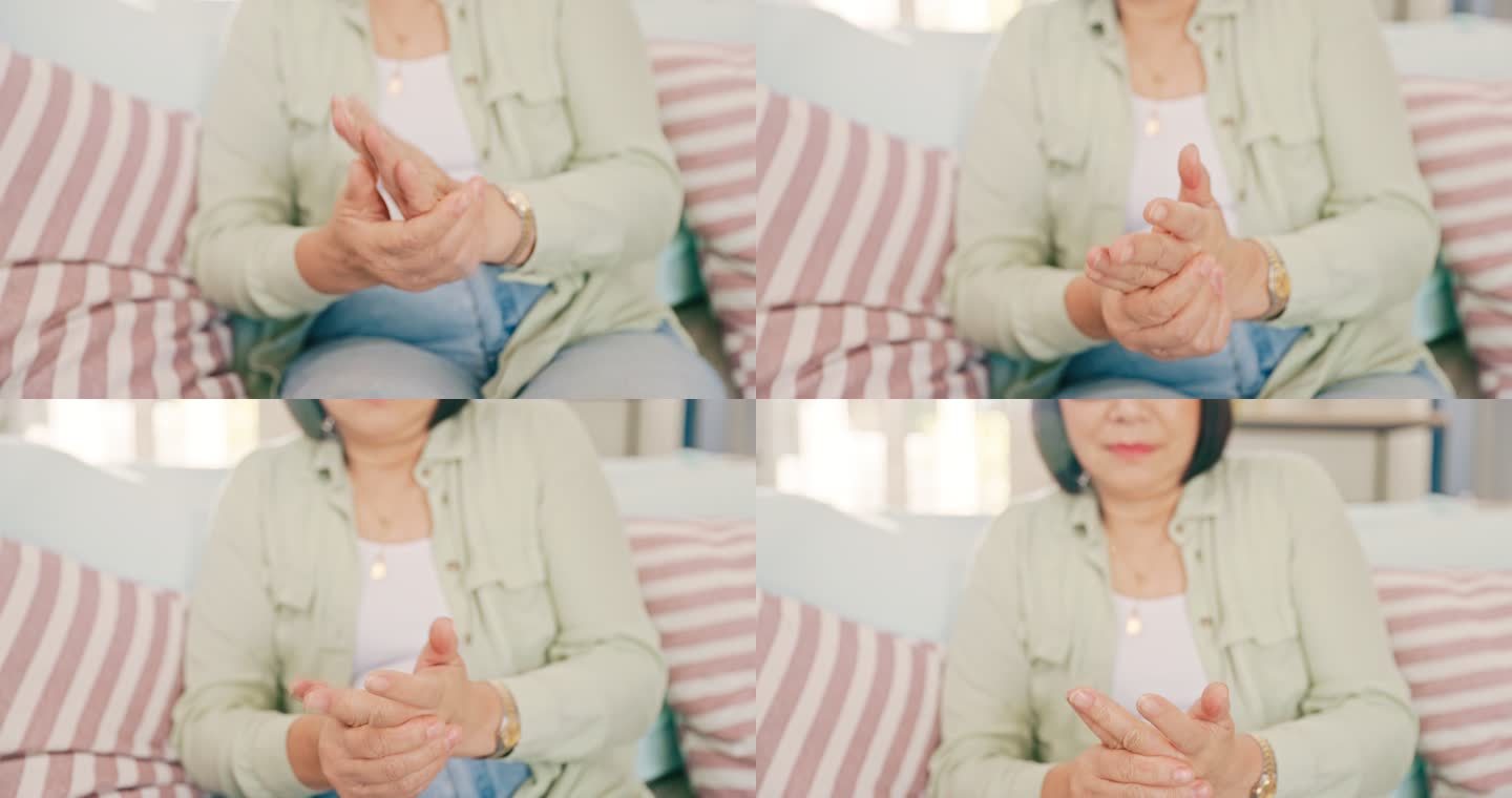 一位亚洲资深女性独自坐在家里的沙发上，双手患有关节炎。一位年长的亚洲妇女在家里的客厅里搓着她酸痛的手