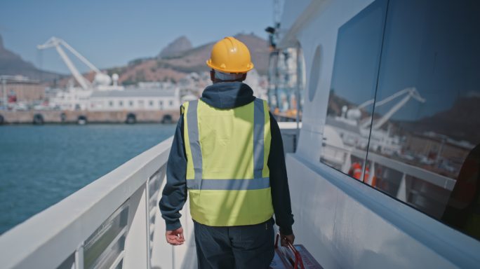 一名修理工戴着安全帽，穿着个人防护装备工作服，手持红色工具箱在货船上行走的后视图。作为机械师在海洋上