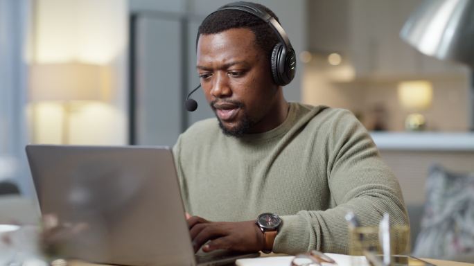 年轻的非洲裔美国呼叫中心特工戴着耳机，在家里的办公室里用笔记本电脑打字。年轻的客户服务代理在使用电脑