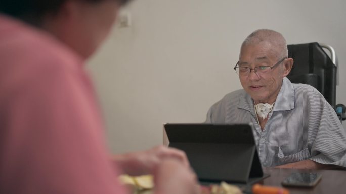 一位坐着轮椅和气管造口管的亚洲老年男子，在晚饭后的闲暇时间，妻子在厨房里把苹果递给他时，他用数码平板