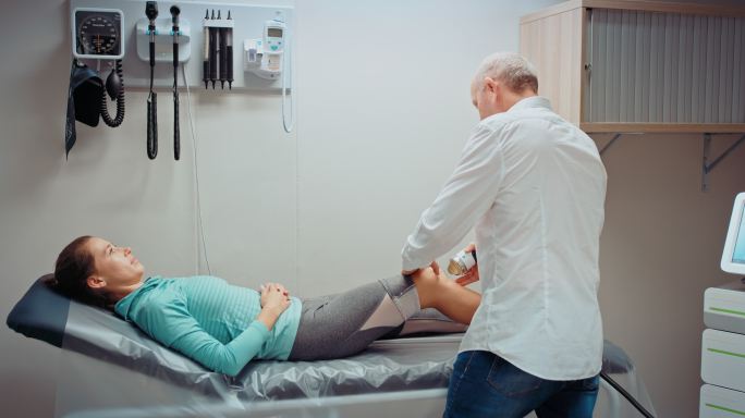 一名医生在一家体育诊所用超声波机检查一名运动女性的腿。物理治疗师帮助患者从膝盖受伤中恢复。运动员在紧