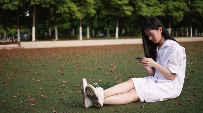 白衣长发年轻美女坐草地上看手机微笑开心