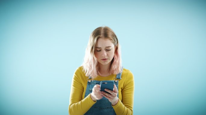 一位女士在蓝色工作室背景下厌恶地看着她的智能手机。一名年轻女性正在使用智能手机，收到坏消息后看起来很