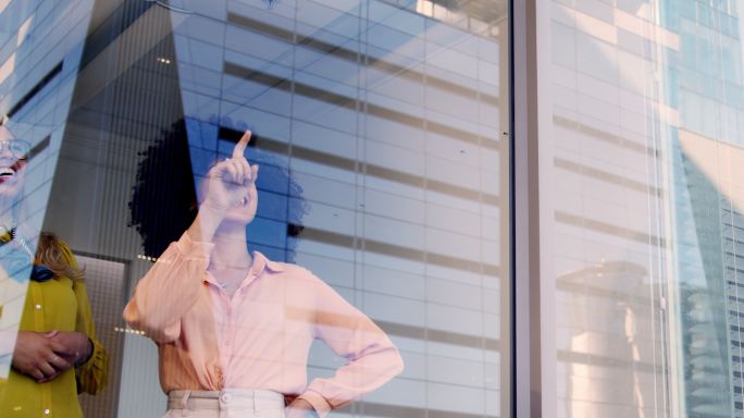 两位年轻的商业女性在现代办公室的玻璃墙上进行头脑风暴。同事们规划和制定战略，在城市背景下指着建筑窗户