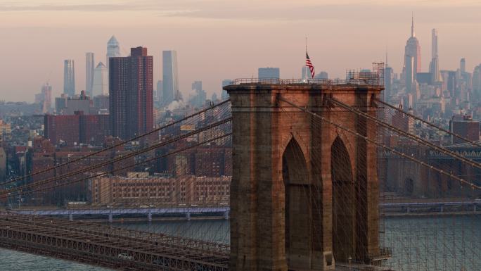布鲁克林大桥上的清晨车流，塔顶挂着美国国旗，背景是曼哈顿天际线的远景，空中视角模糊了。无人机空中视频