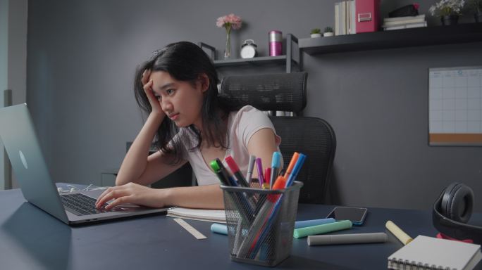 漂亮的亚洲女孩在笔记本电脑上阅读和学习在线课程后感到悲伤和沮丧。家庭学校女孩坐在家里的客厅里担心自己