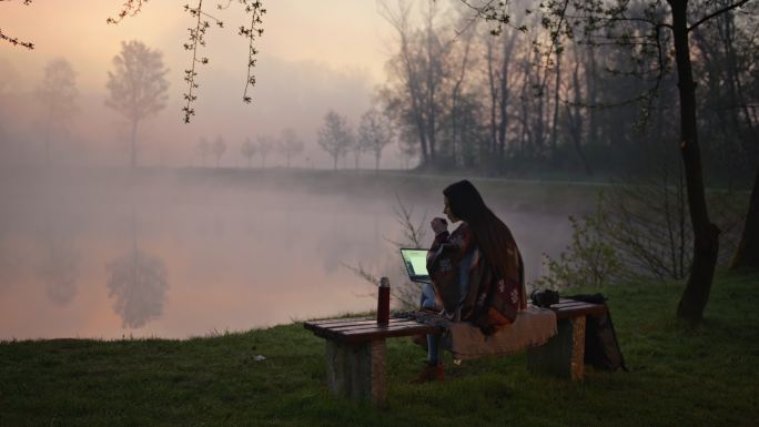 年轻女子坐在雾蒙蒙的湖边的长椅上，一边喝咖啡一边用笔记本电脑工作