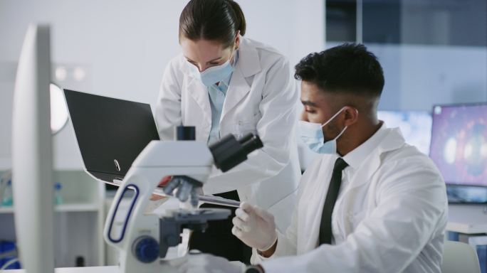 一位西班牙裔科学家在新冠研究设施内观察显微镜。西班牙裔和白种人研究人员分析样本。两位专业人士讨论信息