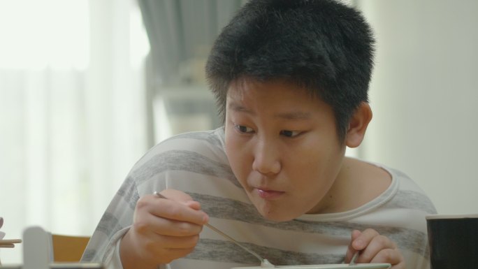 亚洲男孩在家里的餐厅里吃面汤，用智能手机看电影或病毒式视频，这是周末生活方式的概念。