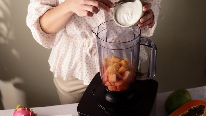 一名妇女在一个装满芒果和木瓜块的搅拌器罐中加入牛奶奶油
