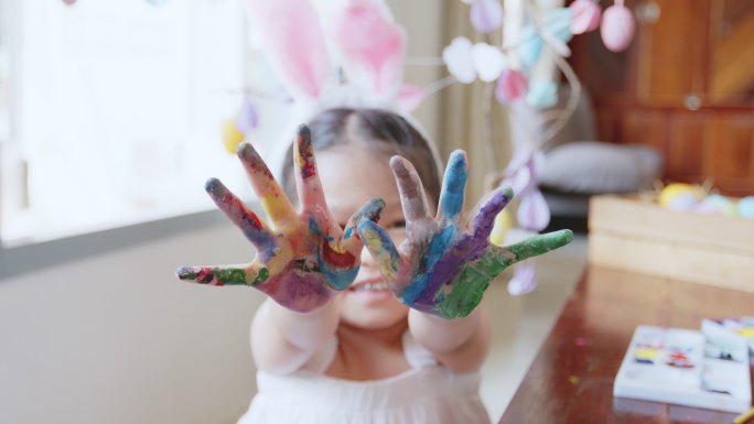 一个可爱的亚洲女孩戴着兔子头巾，用有趣的颜色给她的手着色。孩子们玩得很开心，准备复活节，画彩蛋和装饰