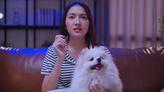 迷人的亚洲年轻美女，晚上在客厅的沙发上和她的狗一起看电视，一边吃爆米花，而她的狗在等宠物，一边吃零食