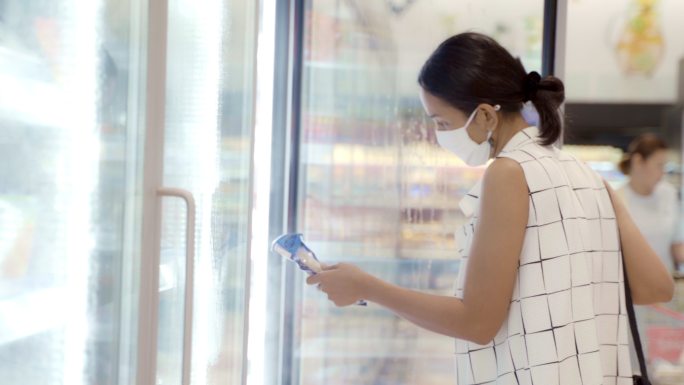 年轻的亚洲女性在超市商店的深冷柜中挑选产品，面罩可防止灰尘和病毒对健康的危害。