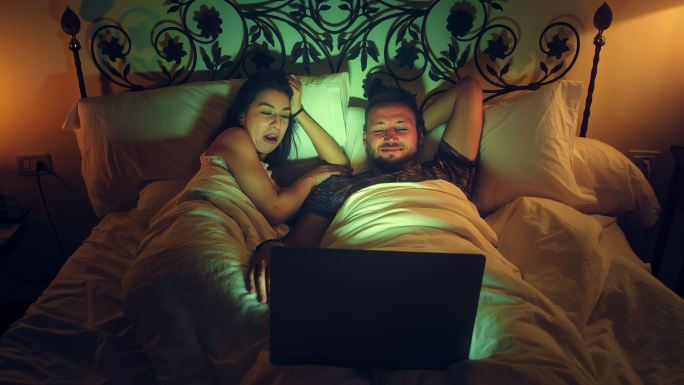 年轻夫妇在床上看电影。使用笔记本电脑并享受乐趣