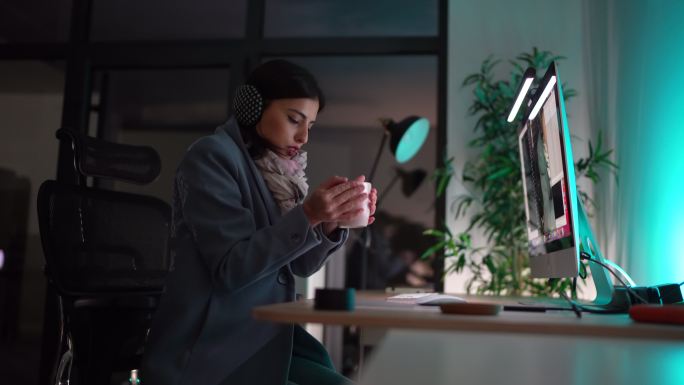 穿着保暖衣服的女人坐在冰冷的办公室里，手里拿着热咖啡杯在电脑上工作