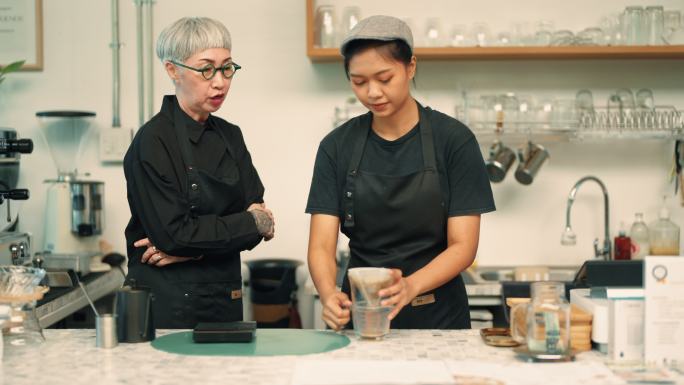 亚洲资深咖啡师企业主正在咖啡馆柜台教一名员工如何制作咖啡。