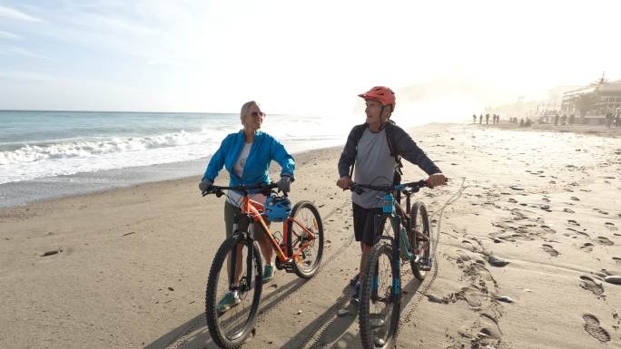 情侣们沿着海滩推着自行车，后面有冲浪