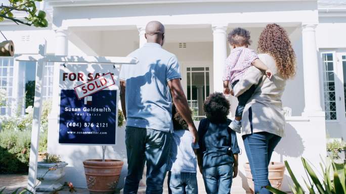 非洲家庭搬进了新家。带着孩子的父母在买了一套新房子后站在前门外，旁边有房产出售的标志