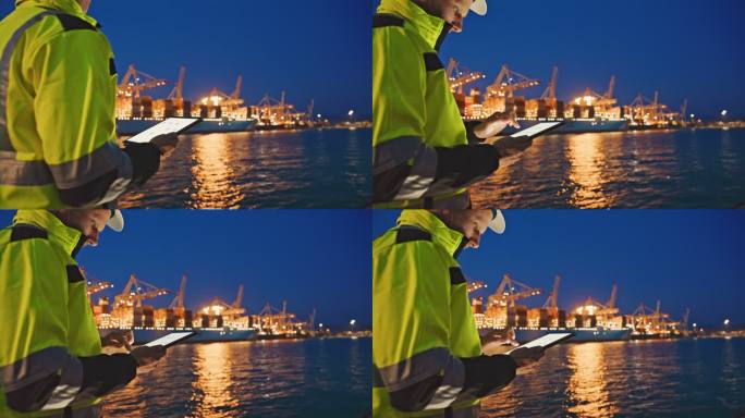 晚上，戴着头盔和警告背心的工程师站在港口一艘大型集装箱船前，使用数字平板电脑