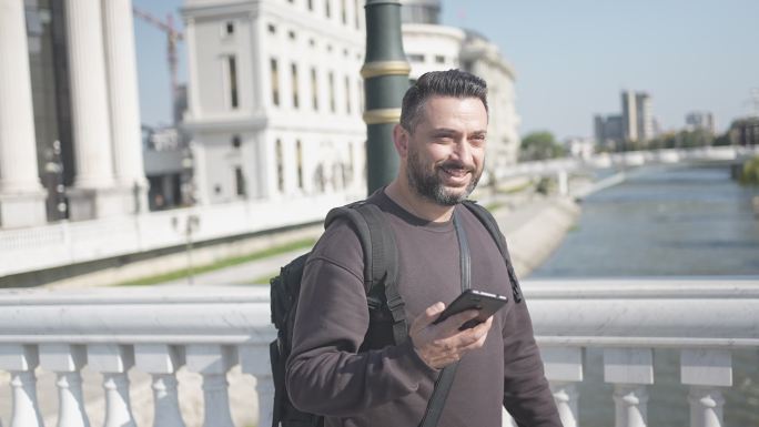 在慢动作视频中，微笑的男性游客在渡河大桥时用智能手机交谈