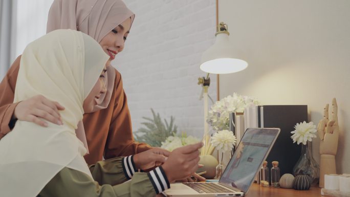 两位身着头巾的穆斯林美女带着信用卡，在家里用笔记本电脑在线订购产品。新的正常购物生活。