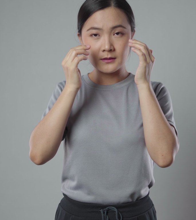 一名亚裔女性因眼睛疼痛而生病，独自站在灰色背景上。垂直视频4k分辨率。