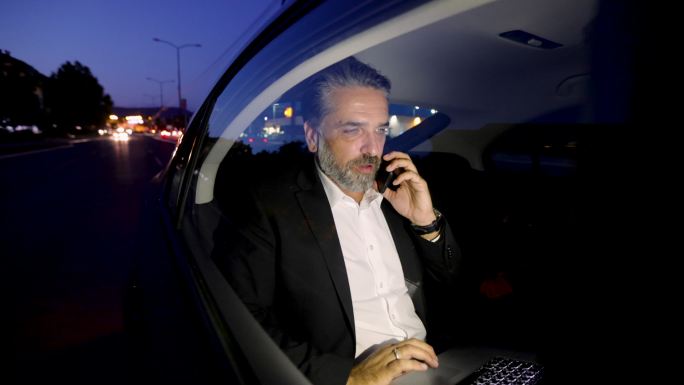 成熟的男人坐在汽车后座上用笔记本电脑和智能手机聊天
