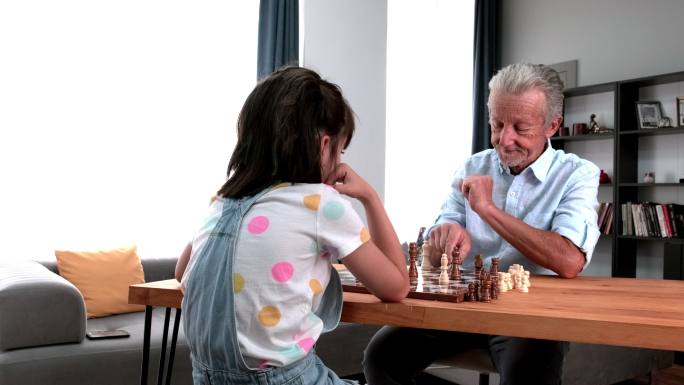 一位开朗的爷爷正和他的小孙女下棋，一起在家度过美好时光